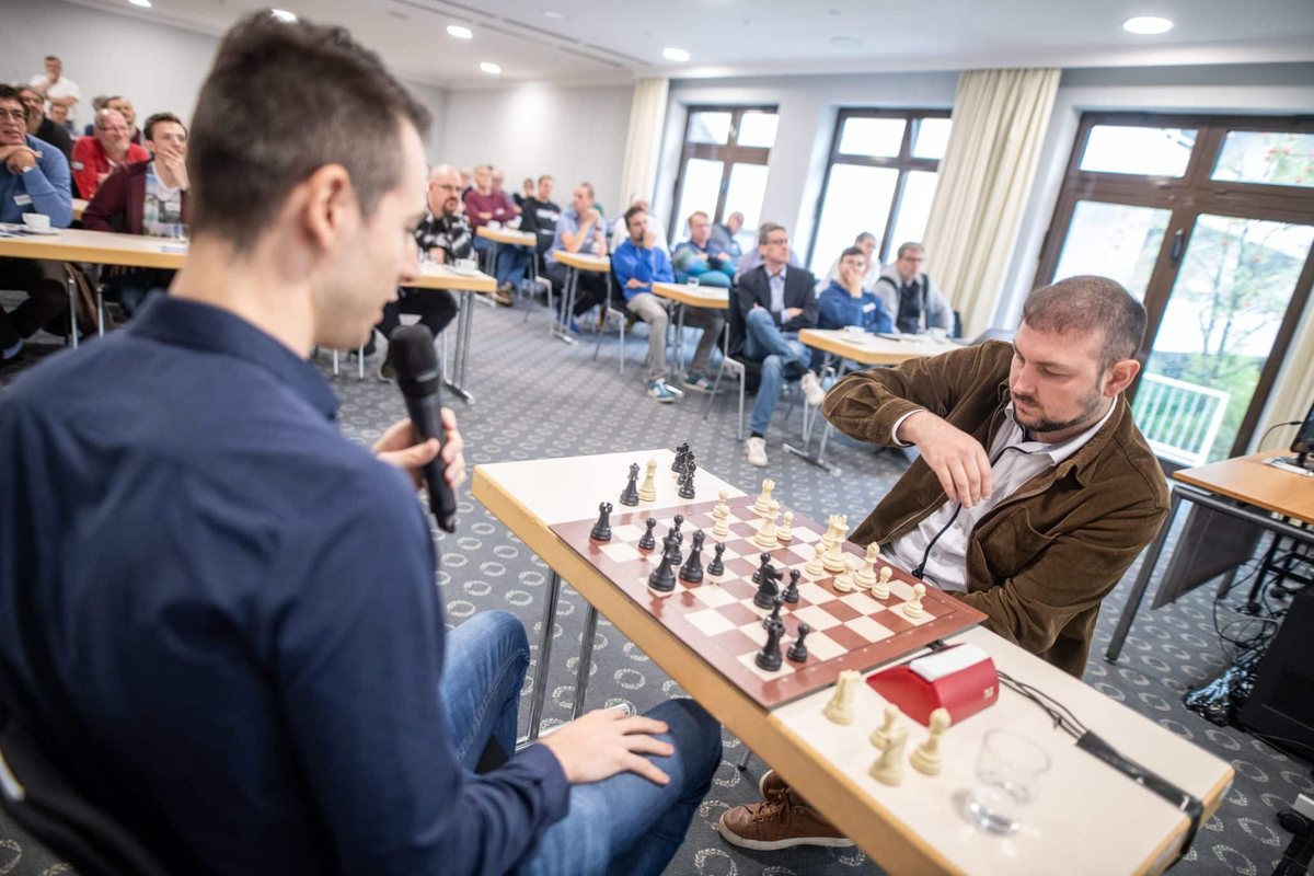 GM Niclas Huschenbeth spielt bei einem Chessence-Seminar vor Ort gegen einen Teilnehmer, im Hintergrund weitere Mitglieder an Schachbrettern